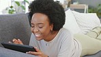 Laughing afro woman using digital tablet to stream funny movies online on subscription channel. Smiling, happy woman lying on home living room sofa, relaxing and using technology for online shopping