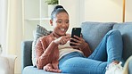 Happy African woman typing on phone while relaxing on her sofa at home. Cheerful black female smiling while browsing internet, chatting on social media or using mobile banking app to track savings