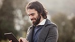 Handsome business man using a tablet and laughing outside on a cold morning. Happy and excited male looking scrolling while standing alone in a park. Trendy young professional guy checking emails 