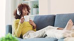 Woman using a phone to text with friends and wearing headphones to listen to music while drinking coffee and relaxing on the couch at home. Cheerful female enjoying songs and the weekend on the sofa