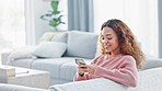 Happy woman texting on a phone while sitting on a sofa inside her living room with copy space. Portrait of a cute young female enjoying online social media, looking comfortable and cheerful at home