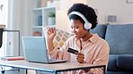 African American female excited about online shopping. Happy woman making payment on her laptop with a credit card. A cheerful lady listening to music with headphones, sitting in front of a sofa.