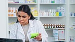 Female pharmacist reviewing medicine, making notes on computer. Young woman is in a pharmacy reading through box of tablets, being serious and recording information on digital screen and keyboard.