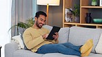 Young man happily laying on couch with tablet. Male is relaxing indoors playing games or using social media for fun. He is isolate and smiling full of joy using digital technology for fun