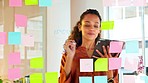 Focused female designer planning ideas on a glass wall with colorful sticky notes inside a modern and creative office. Busy woman enjoying her job while brainstorming projects and managing projects