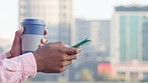 Close up of a hand of an African American person scrolling on phone and holding cup with blurred background of buildings. Web developer texting client. IT professional taking break to drink coffee