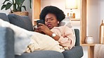 Bored and tired woman lying on a couch typing on her phone at home. A young African female relaxing and yawing on a sofa in her house on a boring weekend. A sleepy lady scrolling social media