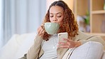 Woman sitting while drinking coffee and on phone alone. Young lady is relaxed indoors on couch at home laughing at social media or texting a friend. She is using technology for entertainment.