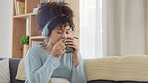 Woman listening to music on headphones or an educational podcast while thinking, drinking a cup of coffee on home living room sofa. Relaxed afro woman enjoying day off and sipping of tea in lockdown