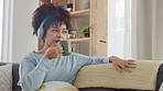 A woman dancing, listening to music on headphones and drinking a cup of coffee while relaxing on a home living room sofa. Cool afro woman enjoying an off day and sipping a mug of tea during lockdown