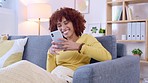 Happy black female texting on a phone while sitting on a couch at home. African American woman laughing while chatting online on a dating app. Lady reading funny memes or uploads on social media