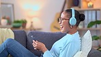 Cheerful female enjoying her favorite song or singer phone using headphones. Happy African American woman listening to karaoke music on a sofa. A smiling lady dancing to a beat or rhythm.