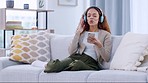 Woman streaming music on her phone while wearing wireless headphones and relaxing at home. Cheerful female sitting on cozy couch enjoying and grooving to favourite songs on an app over the weekend 
