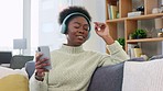 African American female listening to to her favorite song on a phone with headphones. Smiling woman enjoying music while sitting on a sofa. Happy black lady grooving and dancing to a rhythm or beat.