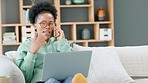 College, university or school student talking on the phone while remote studying from home on her laptop. Young and edgy black woman with afro, wearing glasses and sitting on a living room sofa