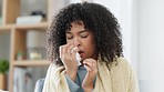 Sick woman with a fever blowing her nose with a tissue while covering herself with a blanket on her couch at home during winter. Young afro girl is feeling ill with a seasonal cold flu or a virus