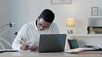 Black man writing notes while working remotely on laptop, analyzing documents in his home office. African American financial analyst checking documents while making a list for a finance report