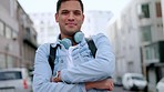 Man with smile and headphones for music or podcast, with arms crossed, in the street, with blurred city background. Young Latino student, with happy expression, has bluetooth earphones and backpack.