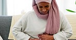 Arab woman, menstruation and stomach pain on the sofa in home living room alone and in agony. Muslim girl taking deep breath while suffering with menstrual period ache in abdomen from uterus.