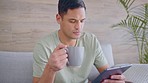 Tablet, coffee and news with a man reading an article online or browsing social media while sitting on his sofa in the living room. Handsome young male surfing the internet to relax in his home