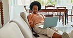 African woman working on laptop on living room couch, noticing smartphone and laughing at phone text. Business communication from home thanks to digital innovation, 5g online internet and mobile tech