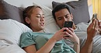 Happy young couple in bed on their phone on social media, browsing the internet and online. Multicultural man and woman laying in bedroom with smartphone. Love, relationship and relax in the morning