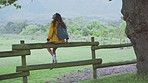 Young woman, backpack and countryside fence for travel while sitting to rest, relax and find peace on a freedom adventure. Backpacker in nature on hiking or trekking journey by rural grass field