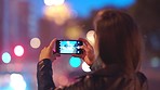 A young woman recording a video of the city at night on her cellphone. A woman using her cellphone to record a video. A young woman out at night in the city using her smartphone