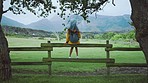 Rain, umbrella and girl on fence in countryside enjoying weather, scenic view and mountain landscape. Wellness, calm and woman sitting on wooden post on farm, field and rural country in spring