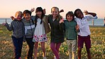 Children group, diversity embrace and smile in park, wave hands and summer by ocean on school trip. Happy multicultural kids, waving and solidarity by sea on lawn for unity, equality or young friends