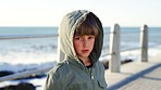 Sad, cold and face of a child at the beach, walking and angry on a holiday at the promenade in Portugal. Unhappy, lonely and portrait of a boy kid at the ocean with a problem during a vacation 