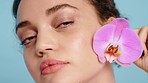 Face, flower and woman in studio for beauty, skincare and nature product, wellness and grooming on blue background. Portrait, girl and model with orchid for facial, eco friendly and floral aesthetic