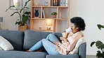 Relaxed Black woman listening to a podcast while drinking coffee and sitting on a couch at home. Happy African American woman streaming online content, enjoying an audiobook while laughing