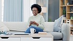 Happy African American woman texting on a phone while relaxing on a sofa and drinking coffee at home. Smiling black woman browsing social media and laughing, planning on how to spend her free time