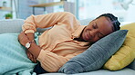 Cramps, stomach ache and woman feeling sick lying on the couch or sofa in a home or house suffering with abdominal pain. Hurt, ill and African America female person touching belly in agony