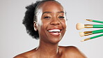 Face, makeup and smile of black woman with brushes in studio isolated on a gray background. Portrait, cosmetics and happy female model laughing with tools to apply foundation or powder for beauty.