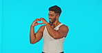 Heart, hand gesture and a laughing man on a blue background in studio for valentines day celebration. Portrait, smile and emoji with a happy young man joking while celebrating love or romance
