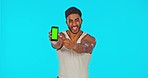 Face, green screen and man with phone pointing in studio isolated on a blue background. Cellphone, thumbs up and happy Indian male flex bicep muscle with advertising, mock up and product placement.