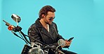 Motorbike, phone and man relax in studio on blue background for road trip, texting and leather aesthetic. Smartphone, motorcycle and man on internet for gps, location and online maps search isolated 