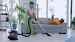 Maintenance, vacuum cleaner and woman cleaning the living room of her house with her husband relaxing. Senior female housewife tidying the floor of dust, dirt and bacteria in her home or apartment.
