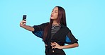 Selfie, fashion and woman with gen z, beauty and confident girl on a blue studio background. Female person, smartphone and model with stylish outfit, trendy and edgy clothes with internet connection