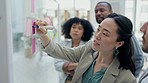 Asian woman, coach and writing on glass board in team brainstorming for schedule planning at office. Female person coaching or training staff on sticky note for strategy or project plan at workplace