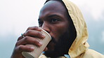 Coffee, morning and black man hiking in the woods with a blurred background of cold, winter weather. Thinking, raincoat and face of a young male hiker in the forest or nature to explore for adventure