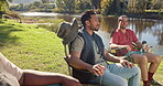 Friends, talking and camping chair in nature, lake or countryside for relax vacation and holiday. Happy, social and group of people or men in conversation for natural carbon footprint by forest river