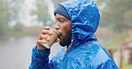 Camping, morning and a black man drinking coffee in the rain while outdoor in nature to relax. Hiking, winter and a young male tourist in the forest or woods for travel and freedom with a cup of tea