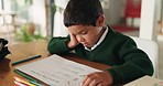 Tired, bored and boy child doing homework, assignment or project in the dining room at home. Burnout, sleepy and little kid student working on school work with books by the table of the modern house.