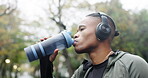 Man, headphones and drinking water in park for sports break, energy and workout. Thirsty african athlete, runner and bottle for nutrition, hydration and recovery of exercise while listening to music 