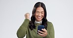 Happy asian woman, phone and winning in celebration, good news or promotion against a grey studio background. Excited female person smile on mobile smartphone app in happiness, sale or discount