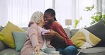 Happy, gift and woman with senior patient on a sofa in the living room for relaxing and bonding. Hug, smile and African girl with present box for elderly female person in the lounge at a modern home.