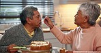 Happy, cake and senior couple in the kitchen eating for tea time and bonding together at home. Love, romance and elderly woman in retirement feeding husband sweet dessert at the dining table at house